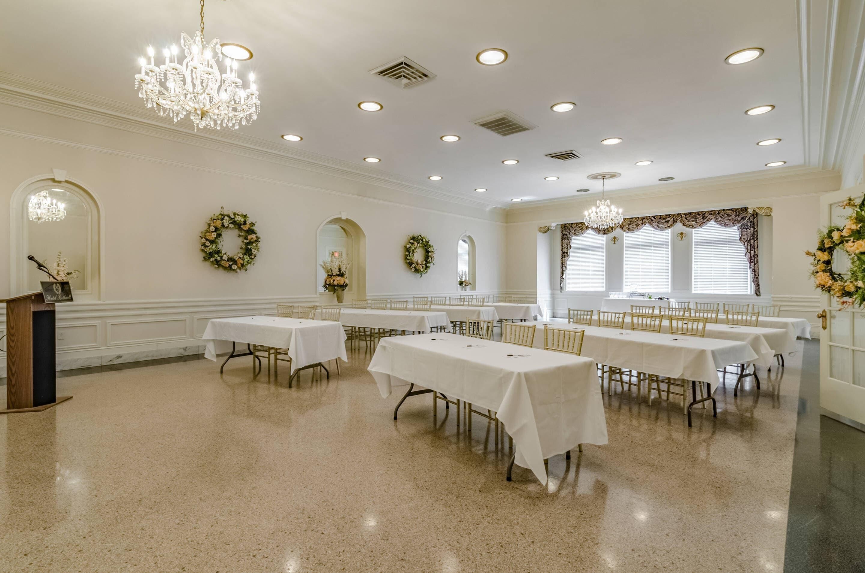 Um salão de banquetes espaçoso com várias mesas, lustres e guirlandas decorativas nas paredes.