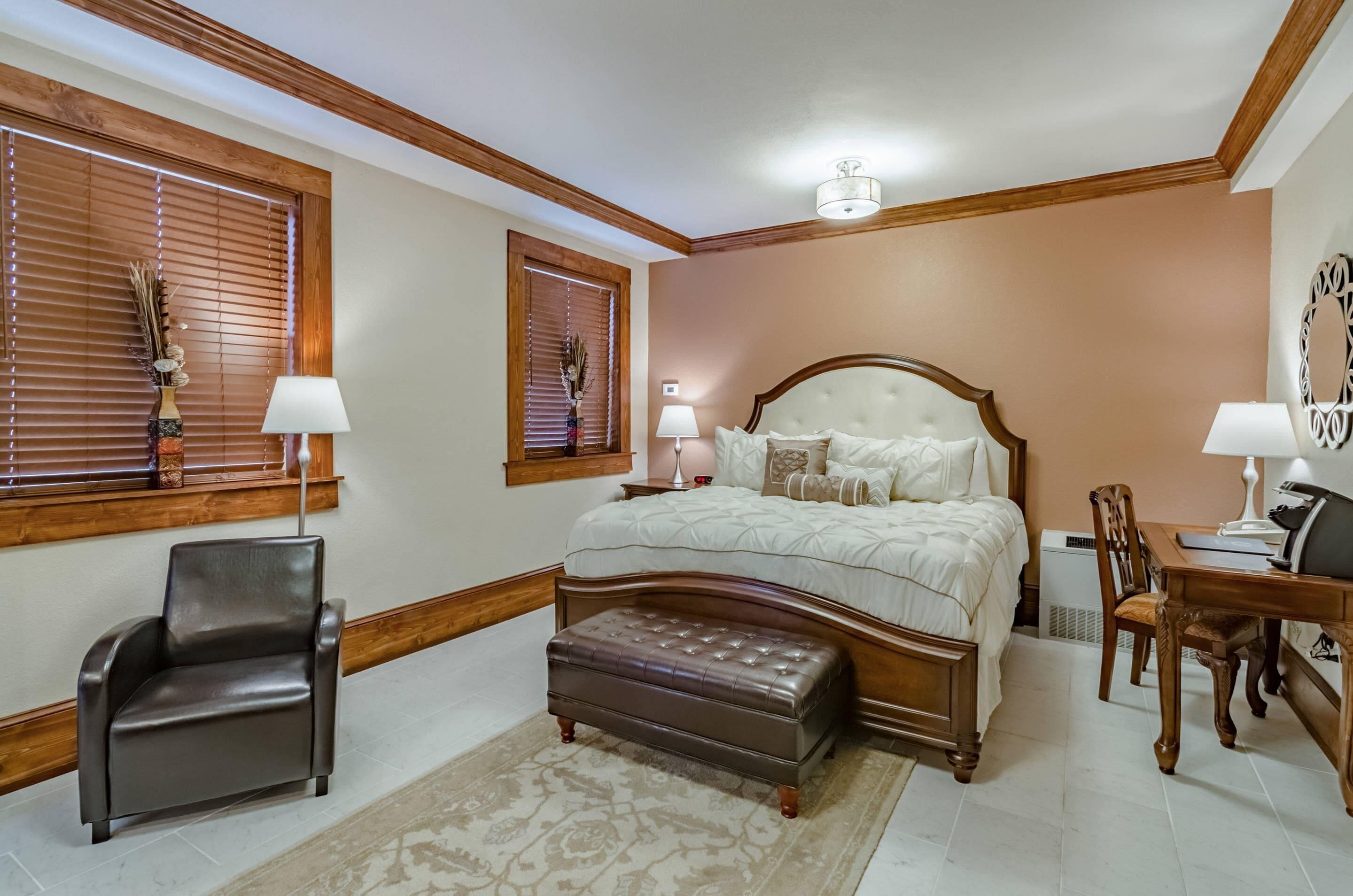 A bedroom with a large bed, wooden headboard, two bedside tables with lamps, a chair, a desk, and a decorative mirror.