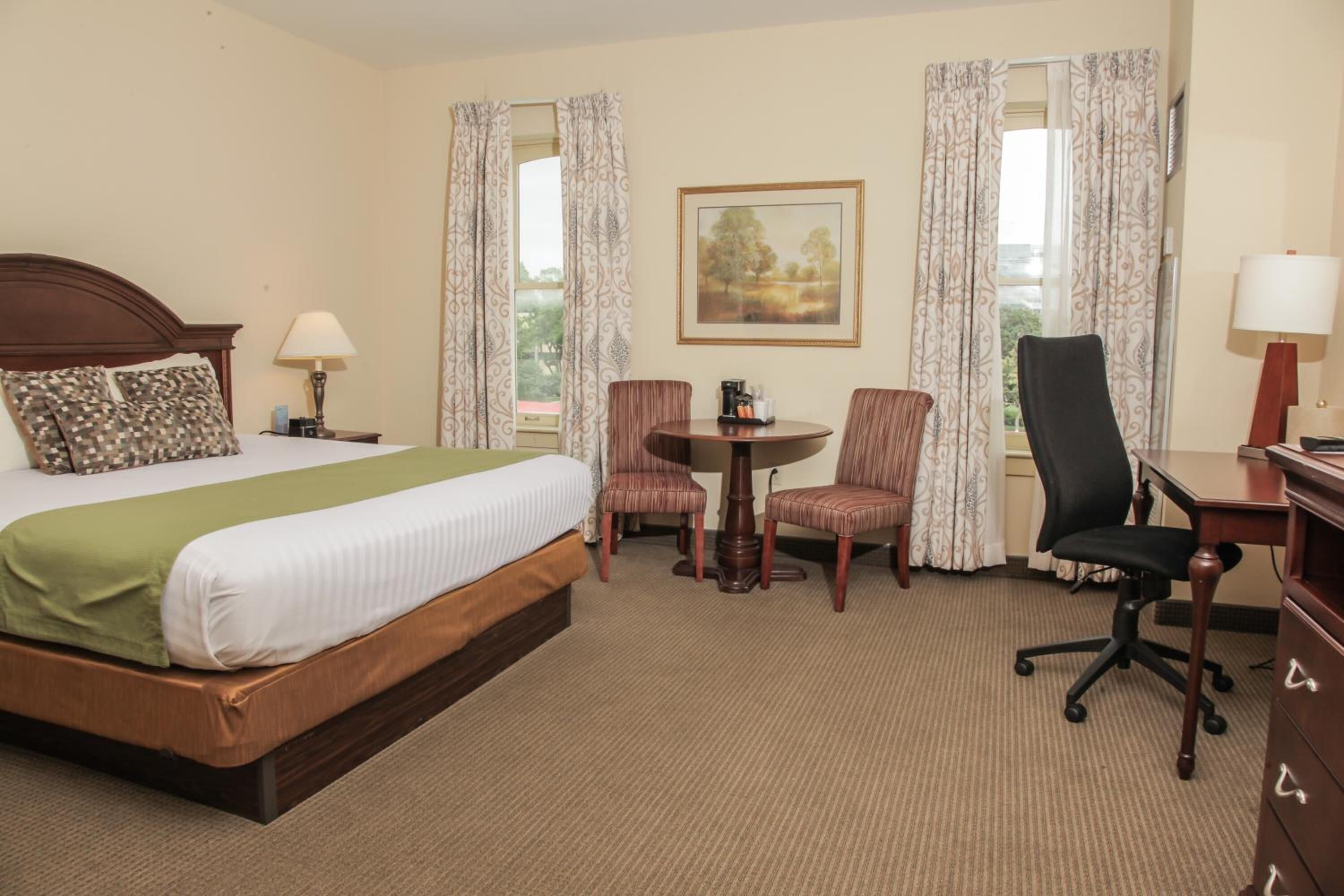 A hotel room with a bed, a desk, two chairs, a table, and a painting on the wall.