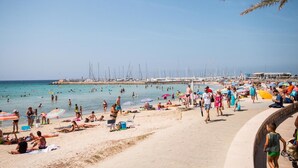 A beach with people sunbathing and swimming, a sandy shore, and sailboats in the background. - Hotel Amic Gala (Palma de Mallorca)