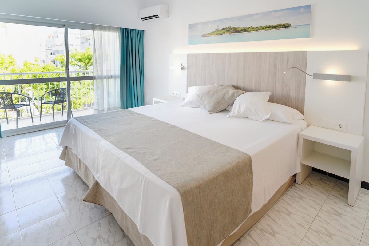 A modern hotel room with a large bed, a balcony with chairs, and a painting of a coastal scene.