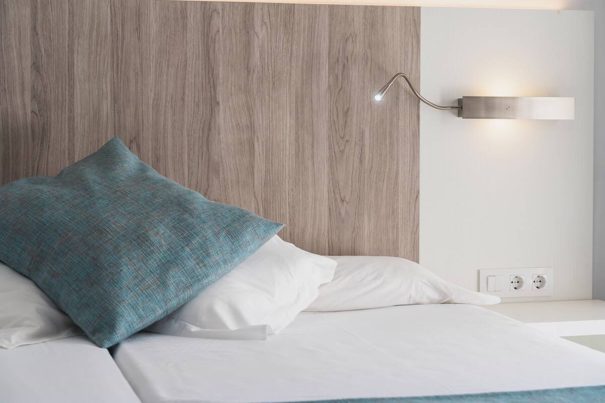 A bed with white bedding and a teal pillow, a wall-mounted light fixture, and a wooden panel wall.
