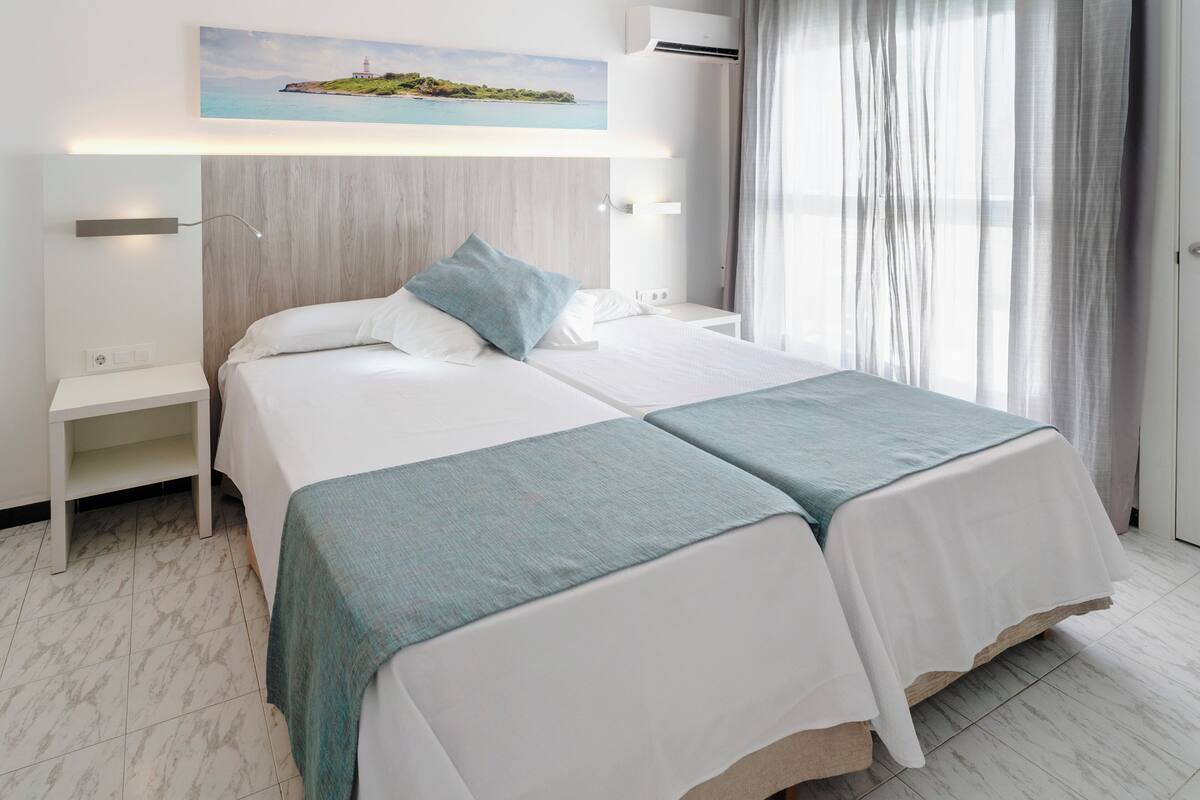 A hotel room with two beds, a nightstand, a wall-mounted air conditioner, and a painting of a coastal landscape.