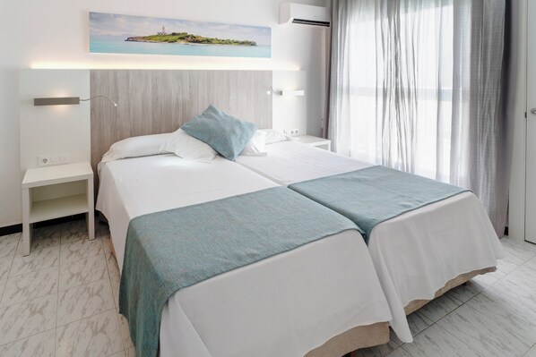 Habitación de hotel con dos camas, una mesita de noche, aire acondicionado de pared y un cuadro con un paisaje costero.