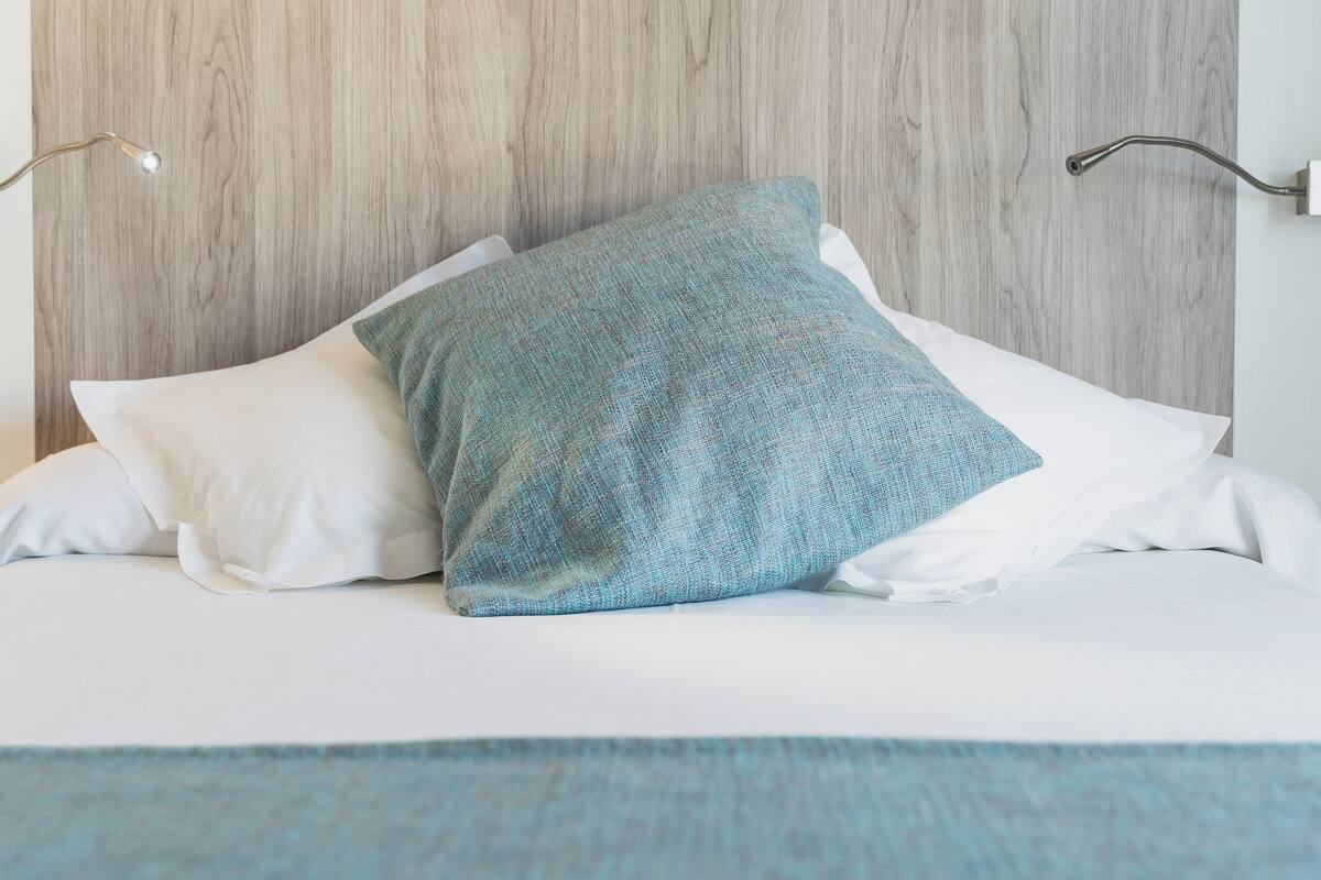 A bed with white bedding and a blue pillow, a wooden headboard, and a wall-mounted lamp.