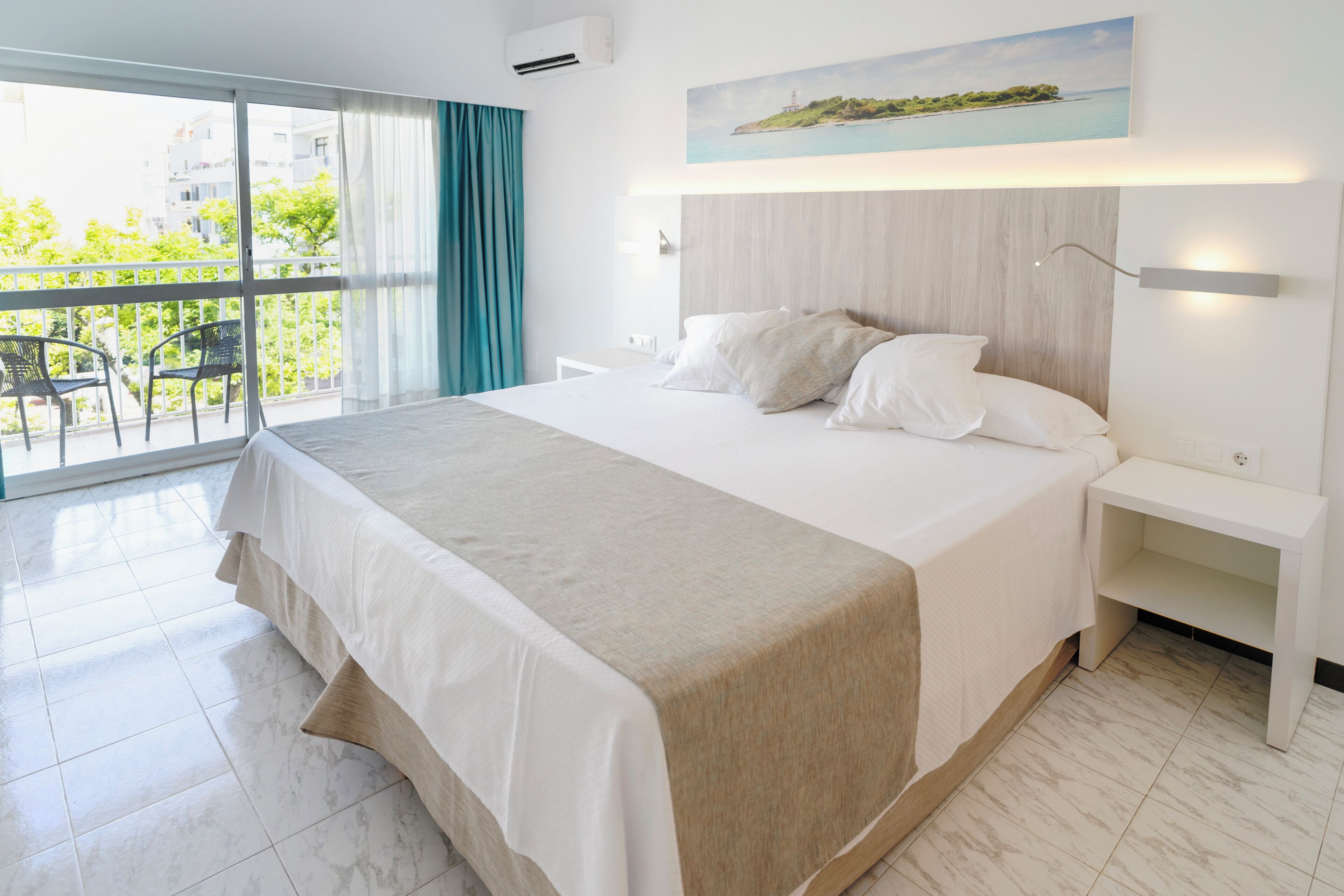 Una habitación de hotel moderna con una cama grande, un balcón con sillas y un cuadro de una escena costera.