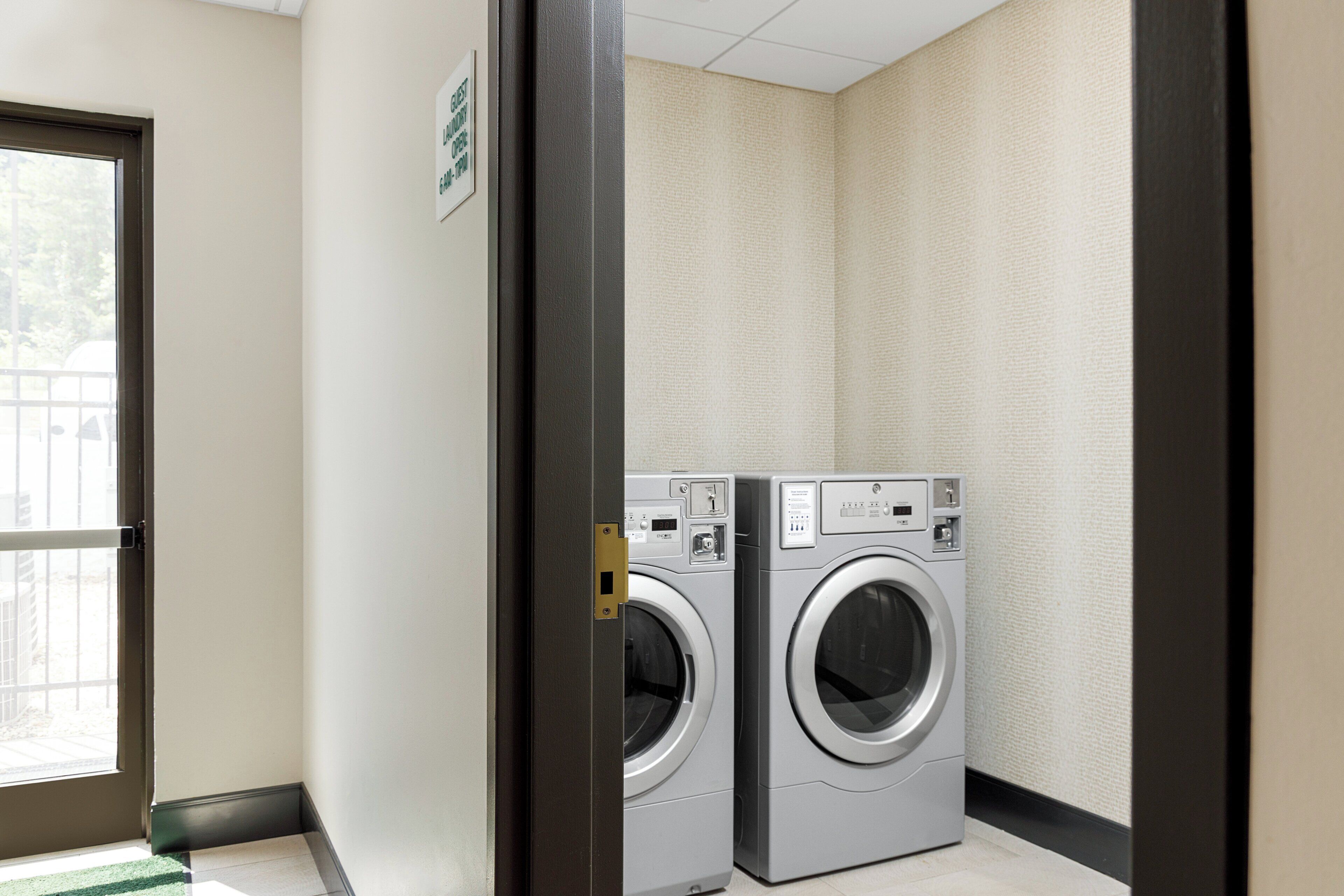 A laundry room with two front-loading washing machines, a door with a sign indicating guest laundry hours, and a window with a view of the outside.