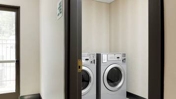 A laundry room with two front-loading washing machines, a door with a sign indicating guest laundry hours, and a window with a view of the outside.