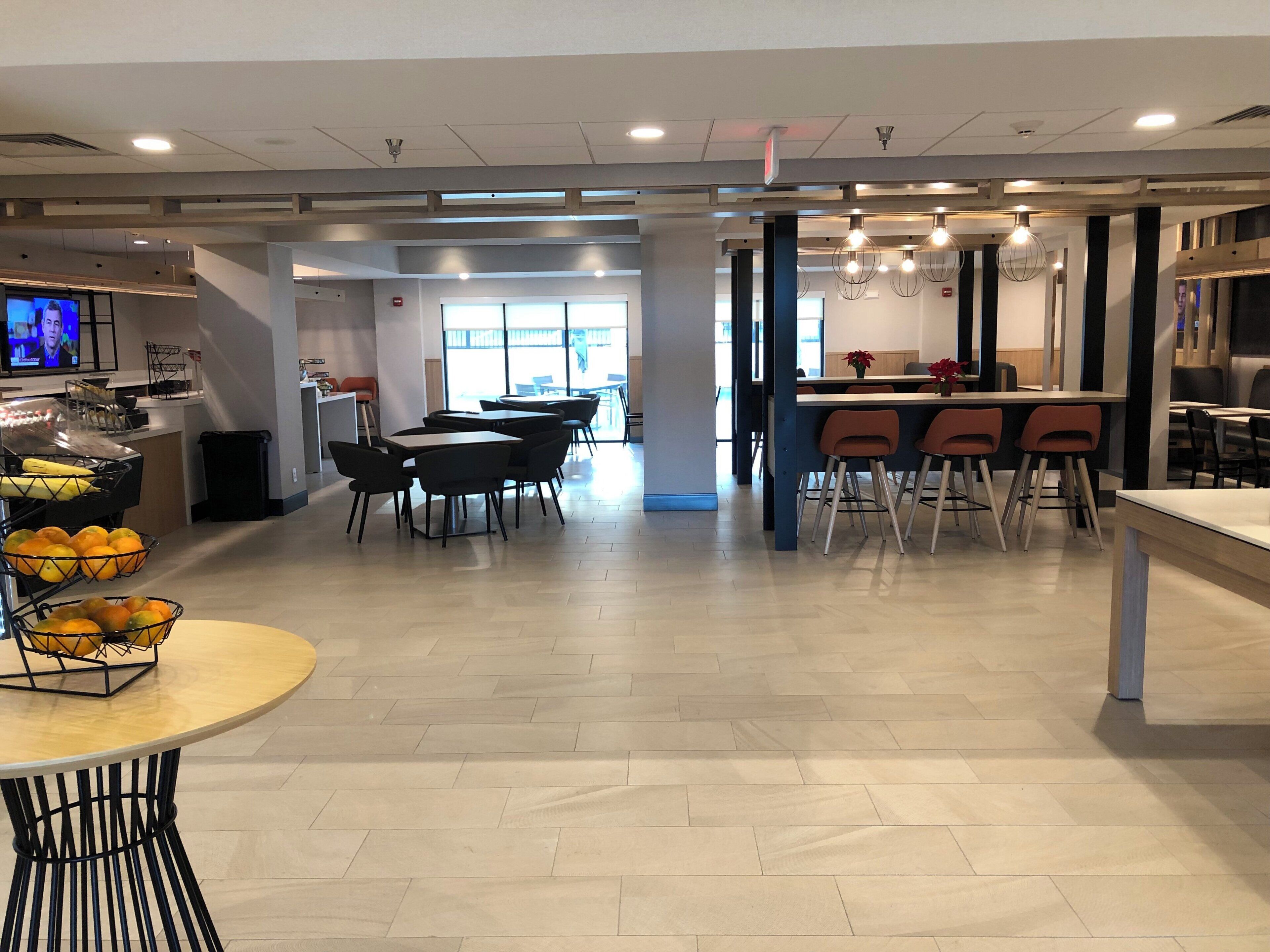 A modern dining area with multiple tables and chairs, a kitchen area with a counter, and a television screen displaying a program.