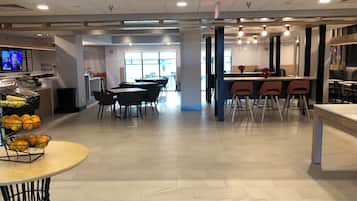 A modern dining area with multiple tables and chairs, a kitchen area with a counter, and a television screen displaying a program.