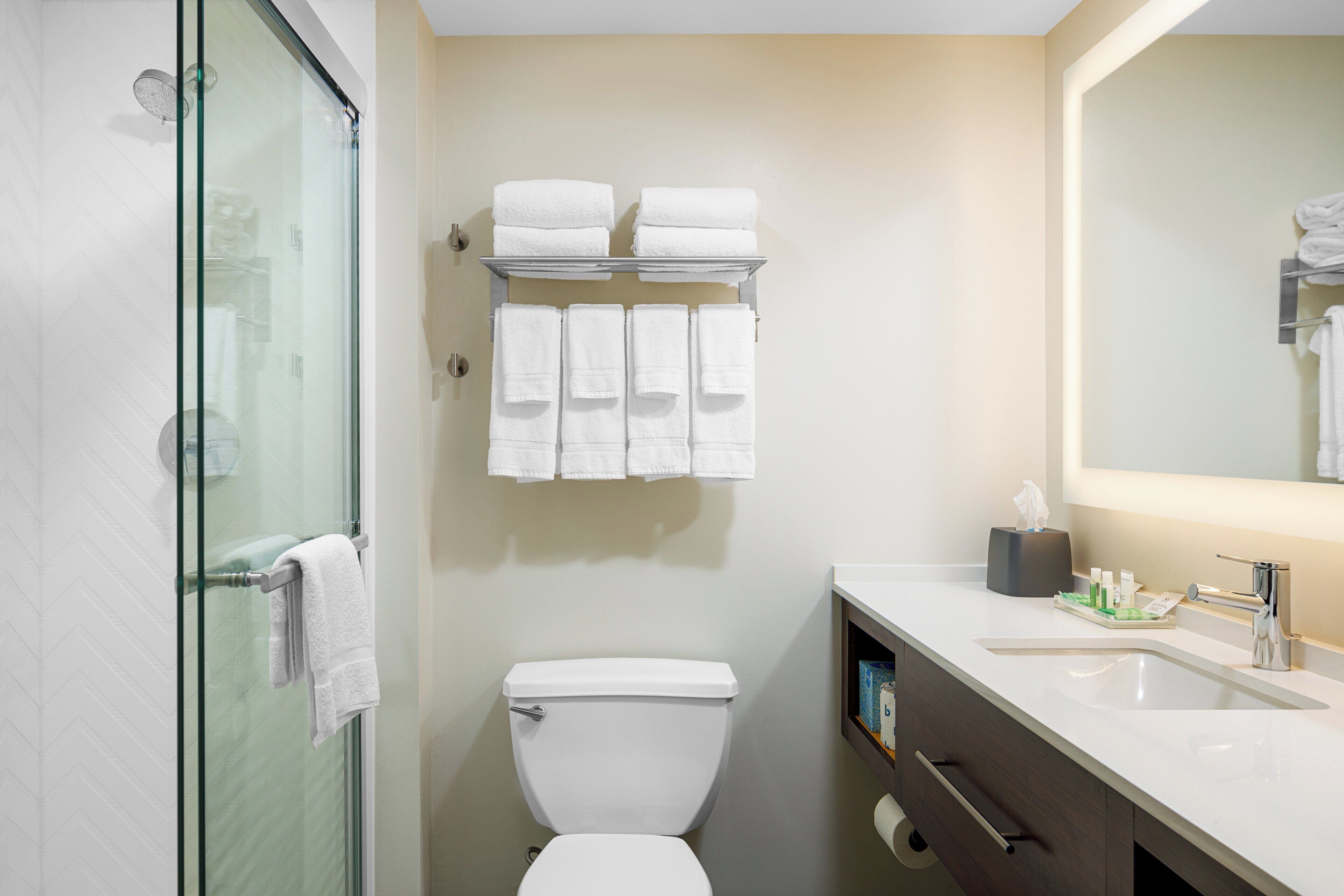 Un baño con cabina de ducha de vidrio, inodoro, lavabo con espejo y toallero con varias toallas.