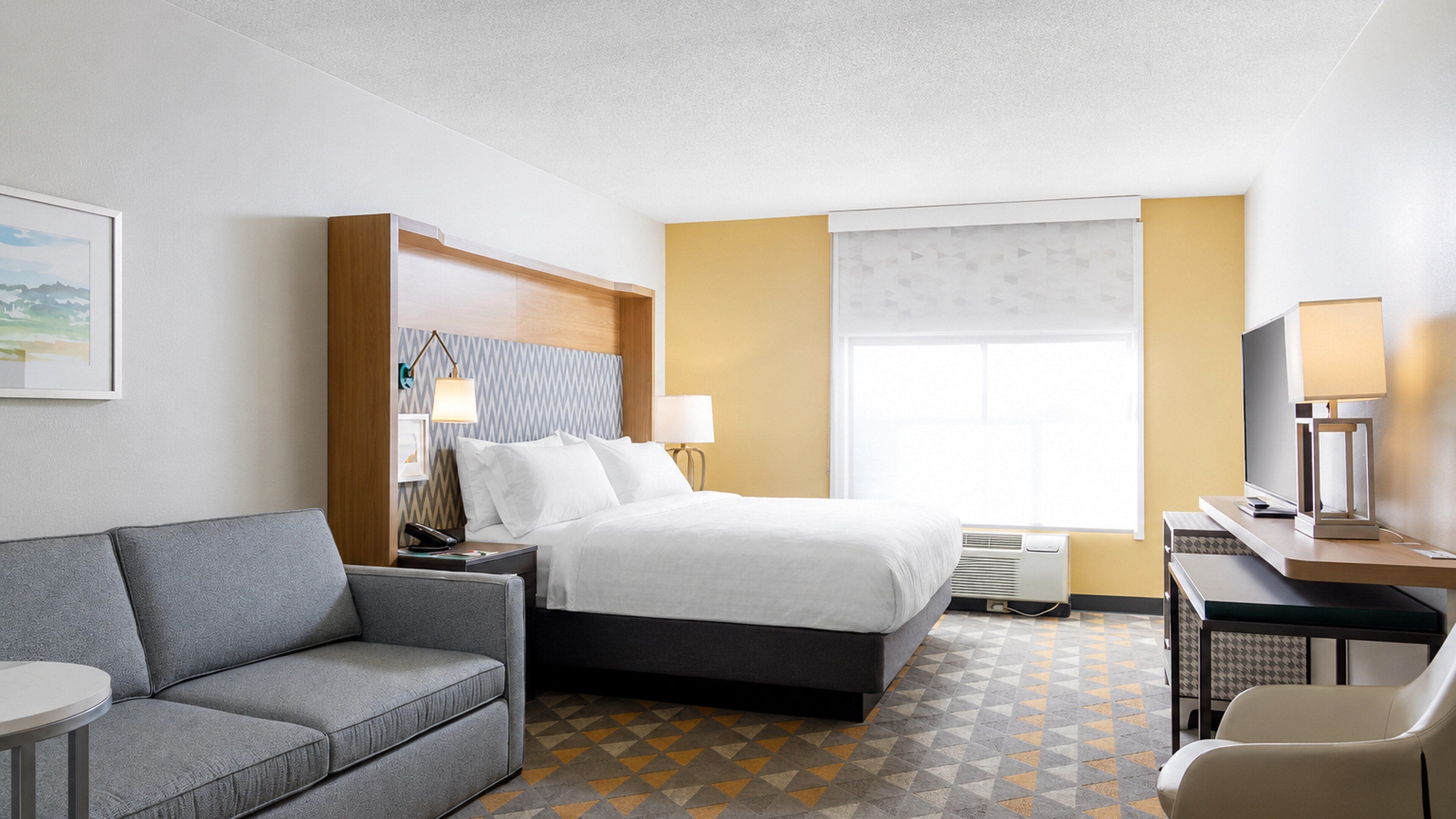Habitación de hotel con cama, sofá gris, escritorio con computadora y televisor.