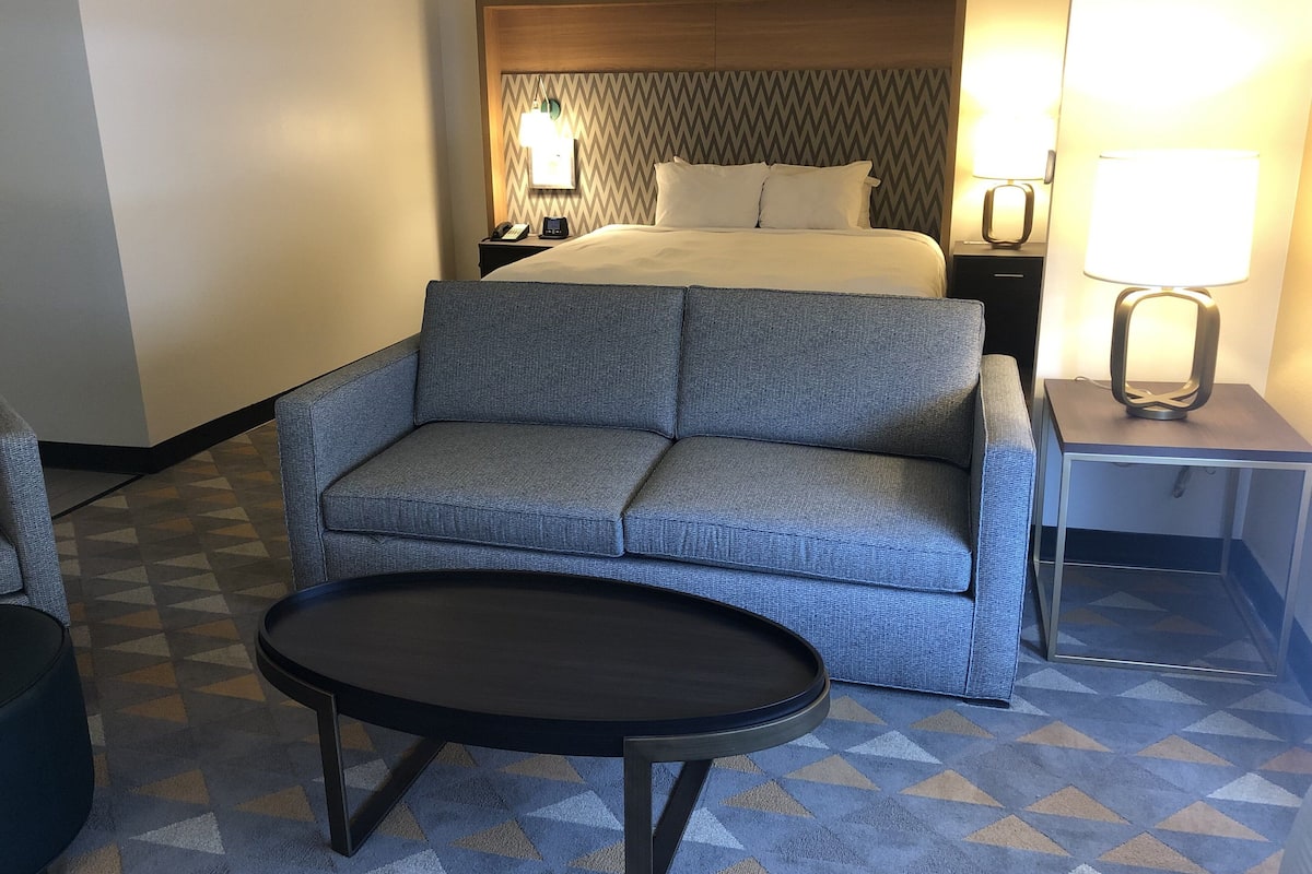 A hotel room with a bed, a grey sofa, a small table, and a nightstand with a lamp.