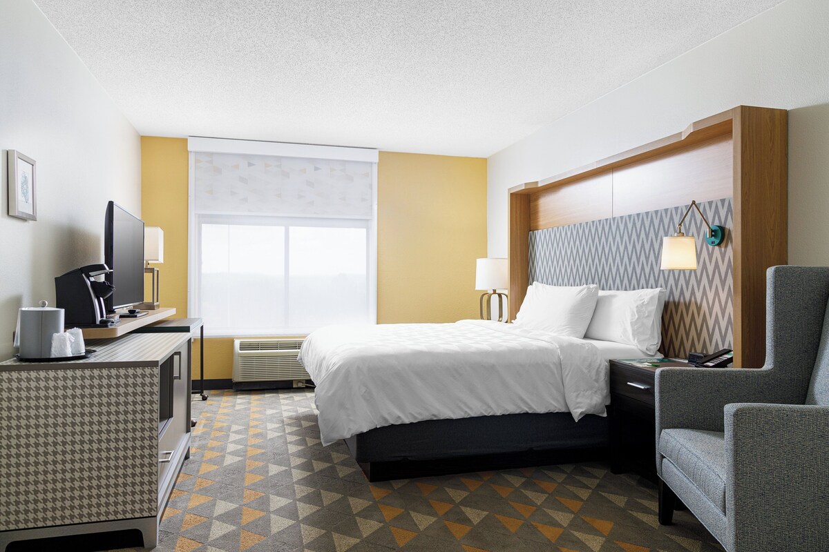 A hotel room with a bed, a desk with a computer, a chair, a TV, and a window with blinds.