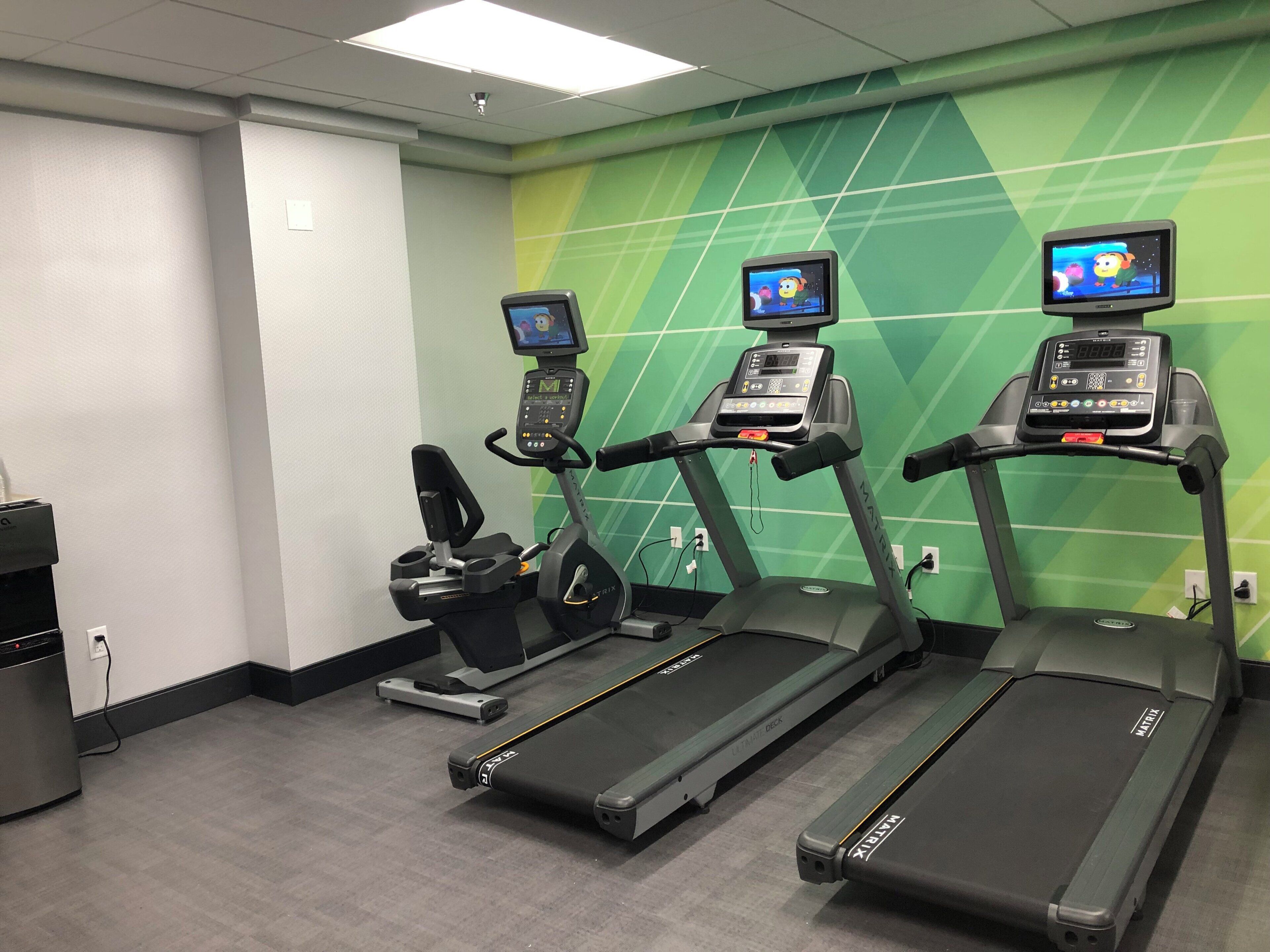 Sala de ginástica equipada com esteiras, bicicleta ergométrica e dispensador de água.