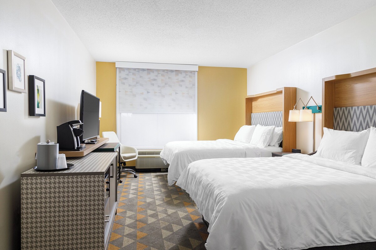 A hotel room with two beds, a desk with a computer, a chair, and a window with blinds.
