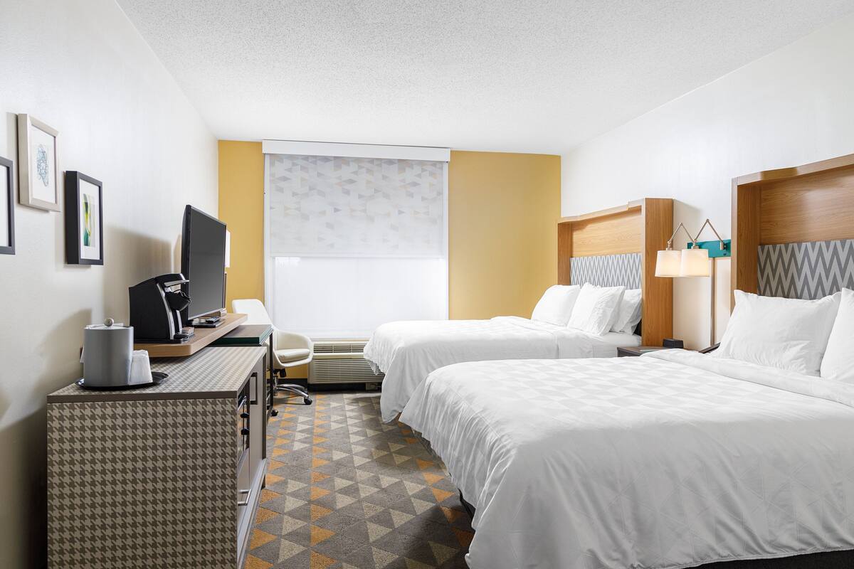 A hotel room with two beds, a desk with a computer, a chair, and a window with blinds.