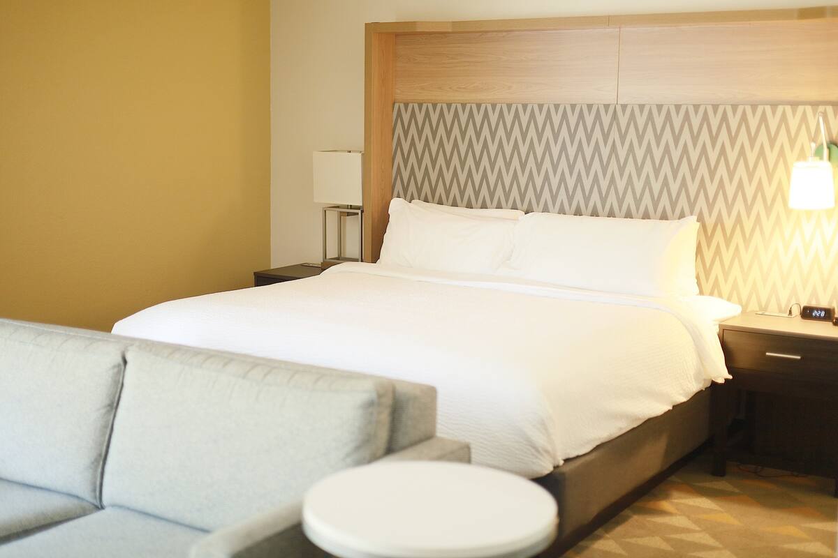 A hotel room with a bed, a nightstand, a lamp, a sofa, and a round table.