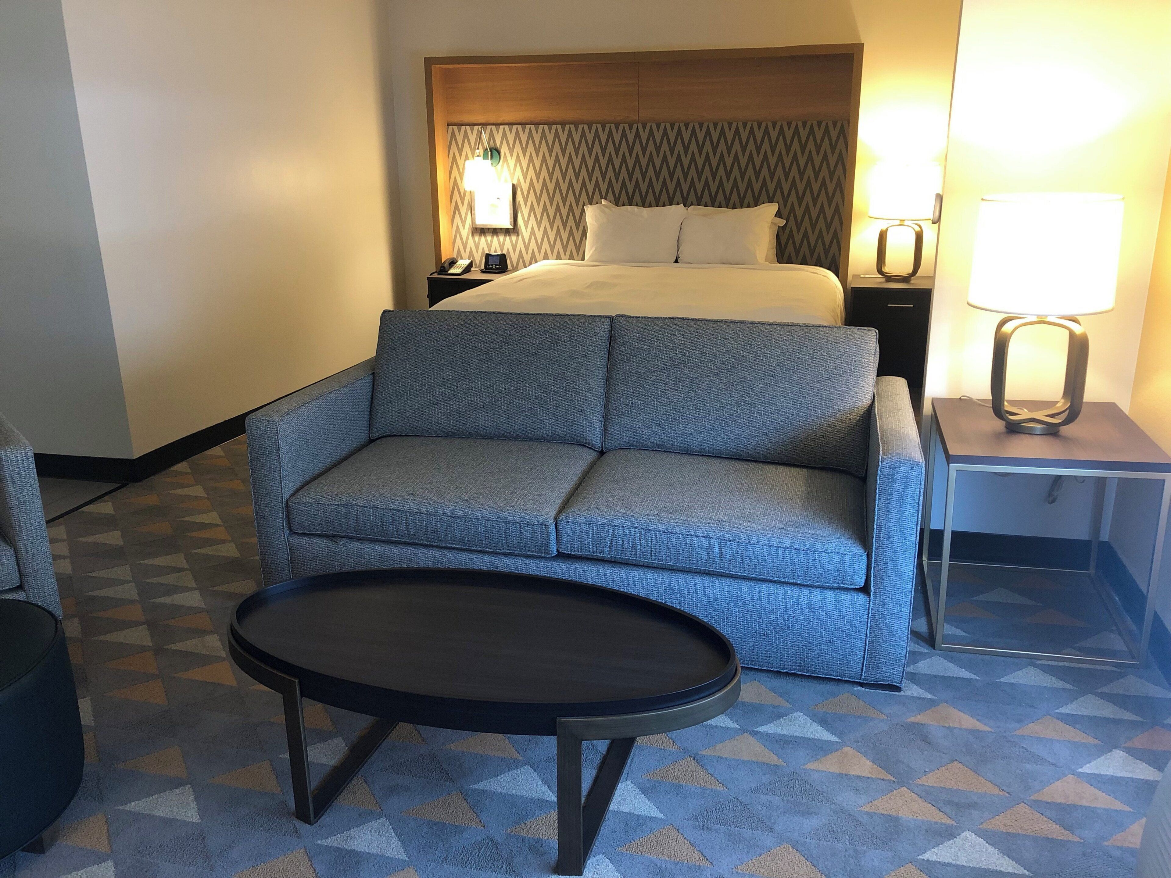 A hotel room with a bed, a sofa, a small table, and a nightstand with a lamp.