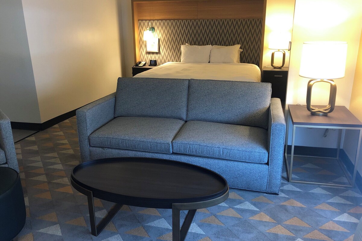 A hotel room with a bed, a grey sofa, a small table, and a nightstand with a lamp.