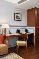A hotel room with a wooden desk, a chair, a lamp, a coffee maker, and a framed picture on the wall.