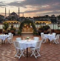 An outdoor dining area with white tables and chairs, a central archway, and a view of a mosque with minarets in the background.