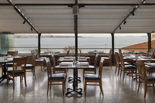 An outdoor dining area with tables and chairs, overlooking a body of water and a distant cityscape.