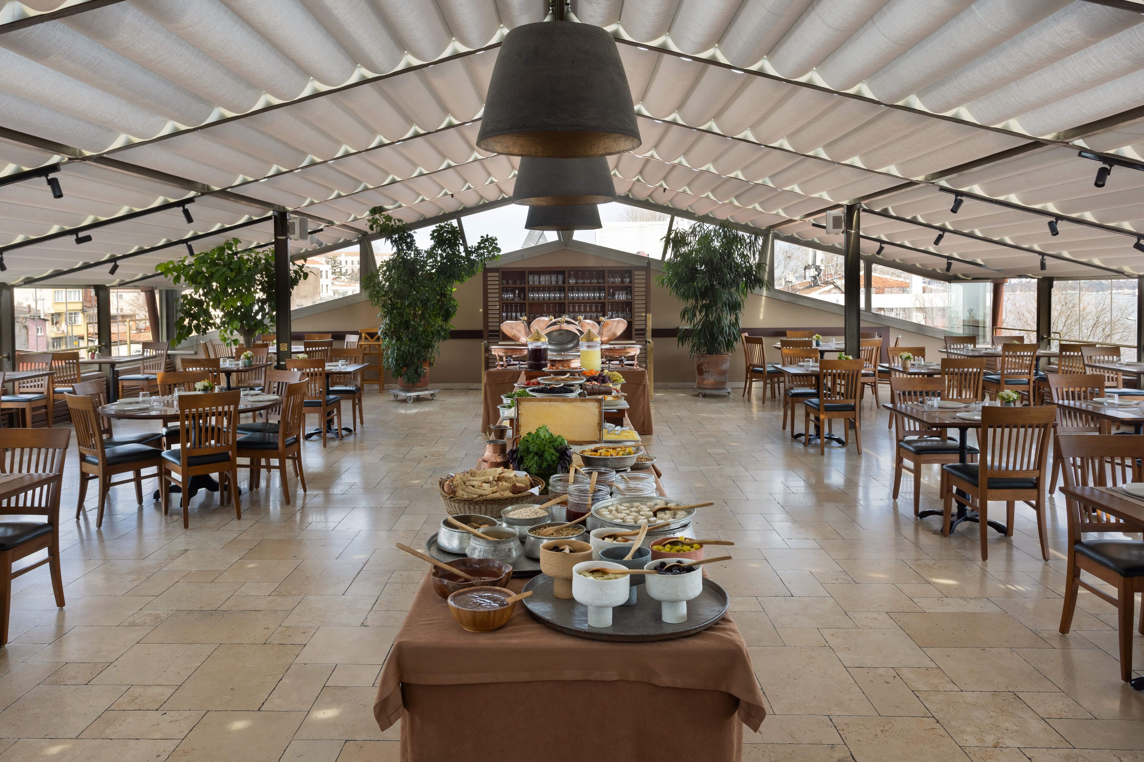 A spacious dining area with multiple tables and chairs, a buffet setup in the center, and large windows providing natural light.