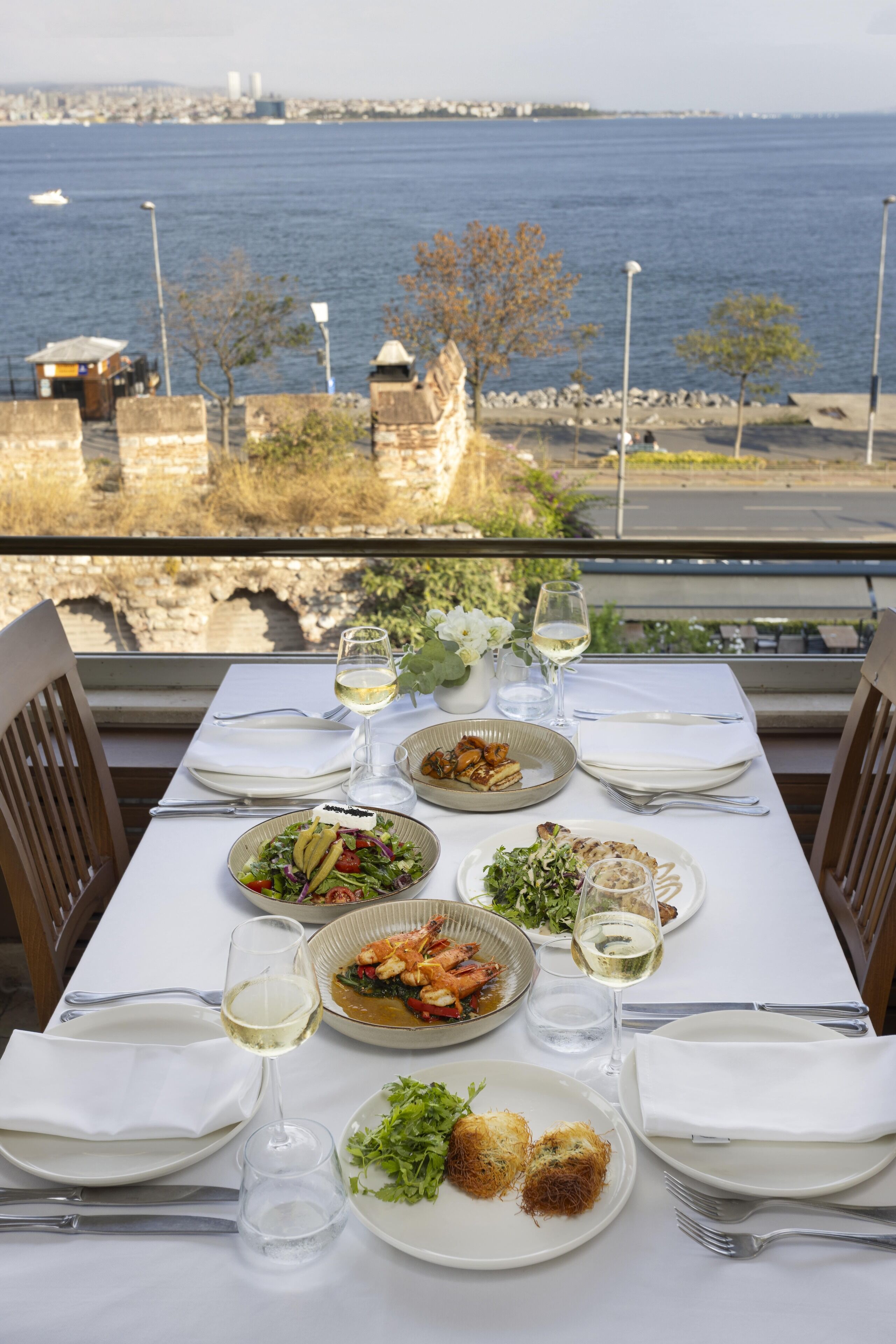 A table set for a meal with various dishes and wine glasses, overlooking a body of water and a historic stone wall.