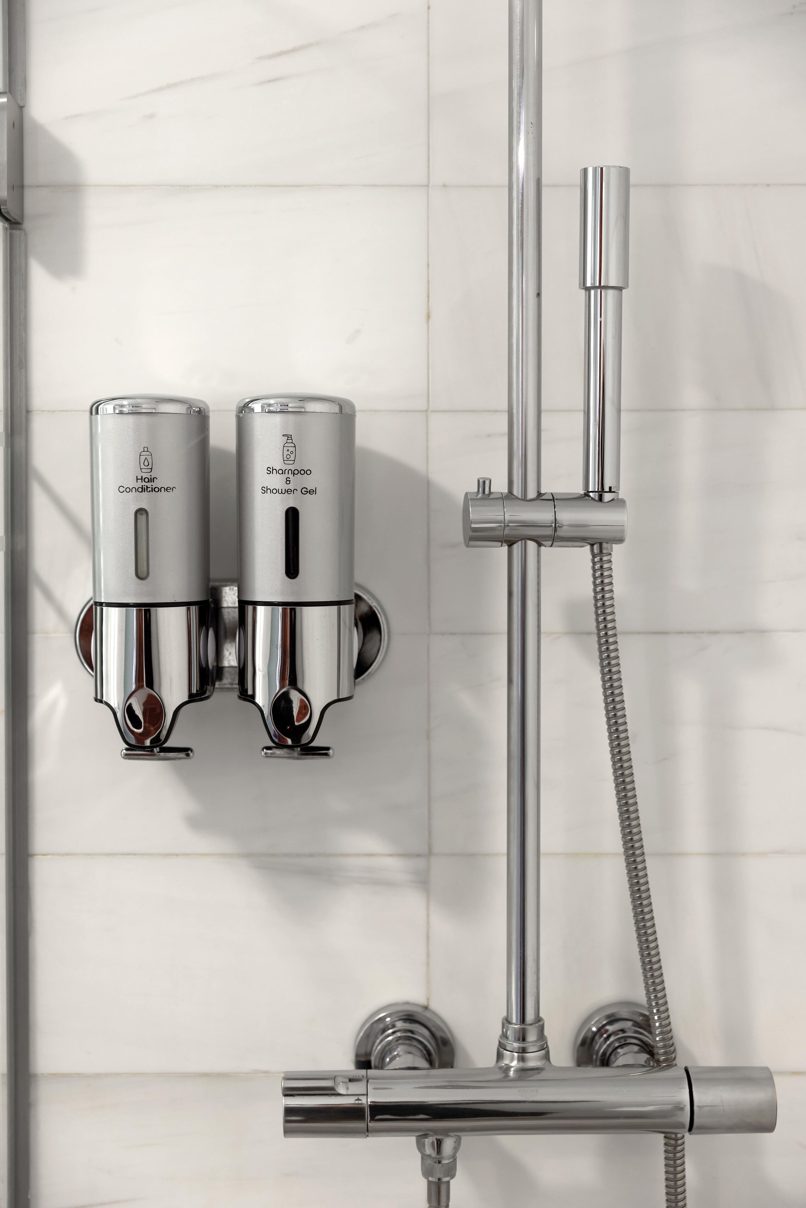 Une salle de bain moderne équipée d’un pommeau de douche chromé, d’un pommeau de douche à main et de deux distributeurs muraux pour l’après-shampoing et le gel douche.