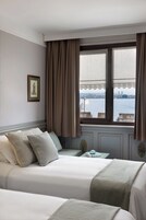 A hotel room with two beds, a window with curtains, a framed picture on the wall, and a view of a body of water and cityscape.