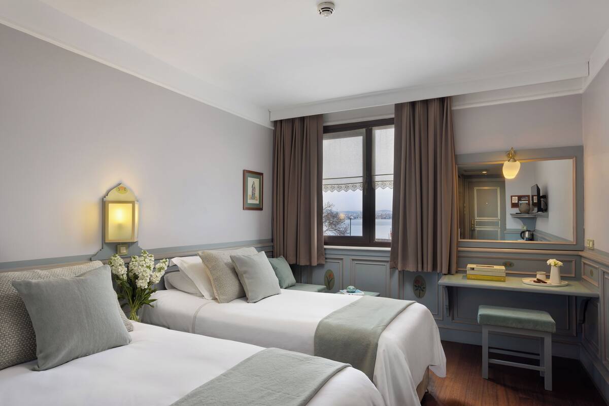 A hotel room with two beds, a desk, a bench, a window with curtains, a wall-mounted lamp, and a mirror.