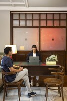 Dos personas sentadas en una mesa de madera con un ordenador portátil, un cartel que dice "Hospitality Desk" y un jarrón con flores.