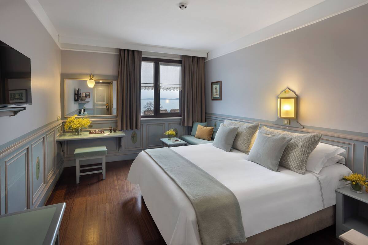 A hotel room with a large bed, a desk with a chair, a small table, a bedside lamp, and a window with curtains.