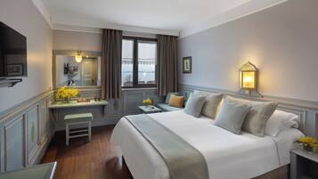 A hotel room with a large bed, a desk with a chair, a small table, a bedside lamp, and a window with curtains.