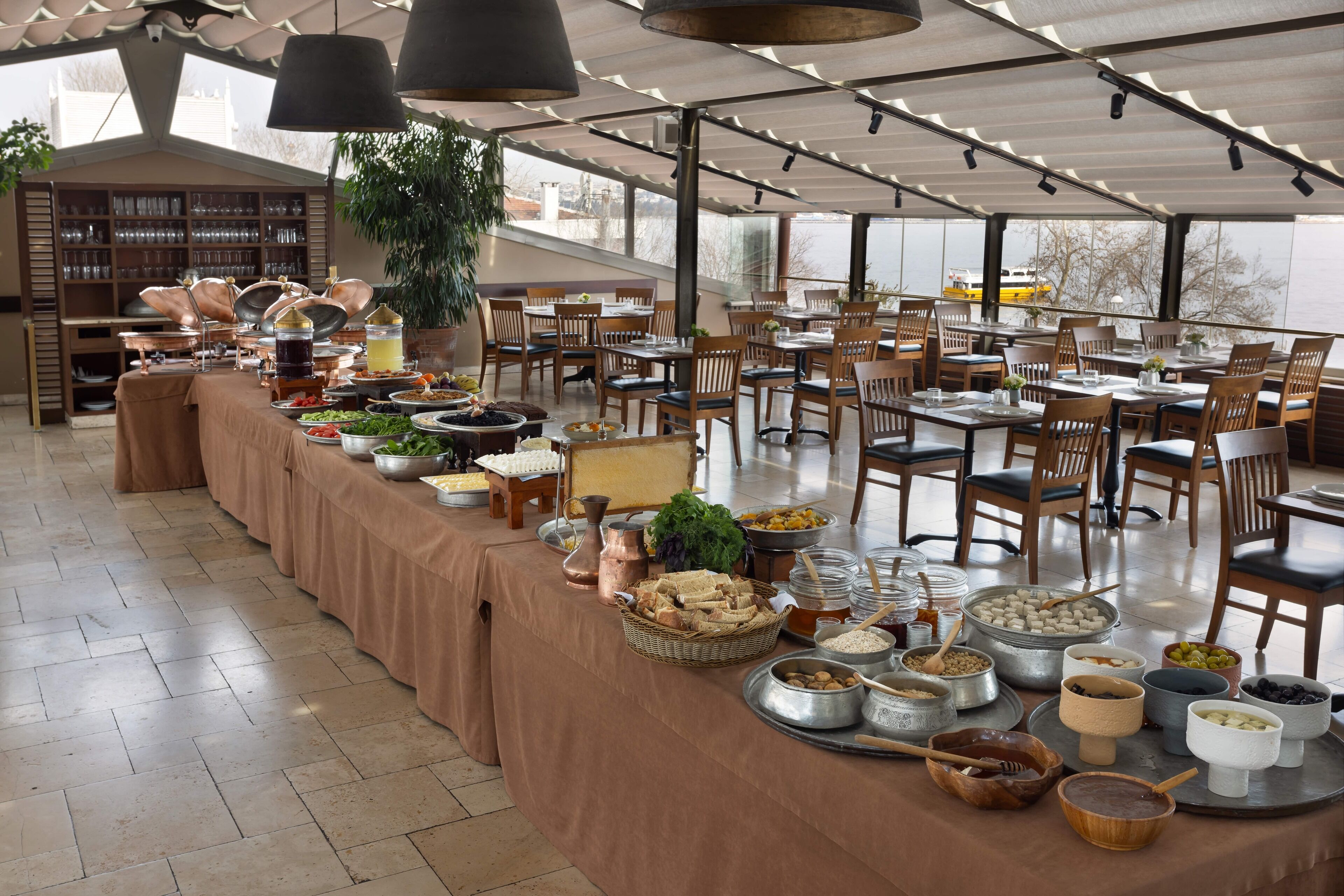 A buffet setup with various food items and utensils on a table, surrounded by chairs and tables in a spacious dining area with large windows.