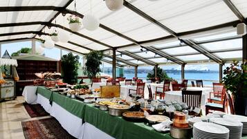 Un área de comedor techada con mesas dispuestas para un bufé, con vista al agua y al horizonte de la ciudad.