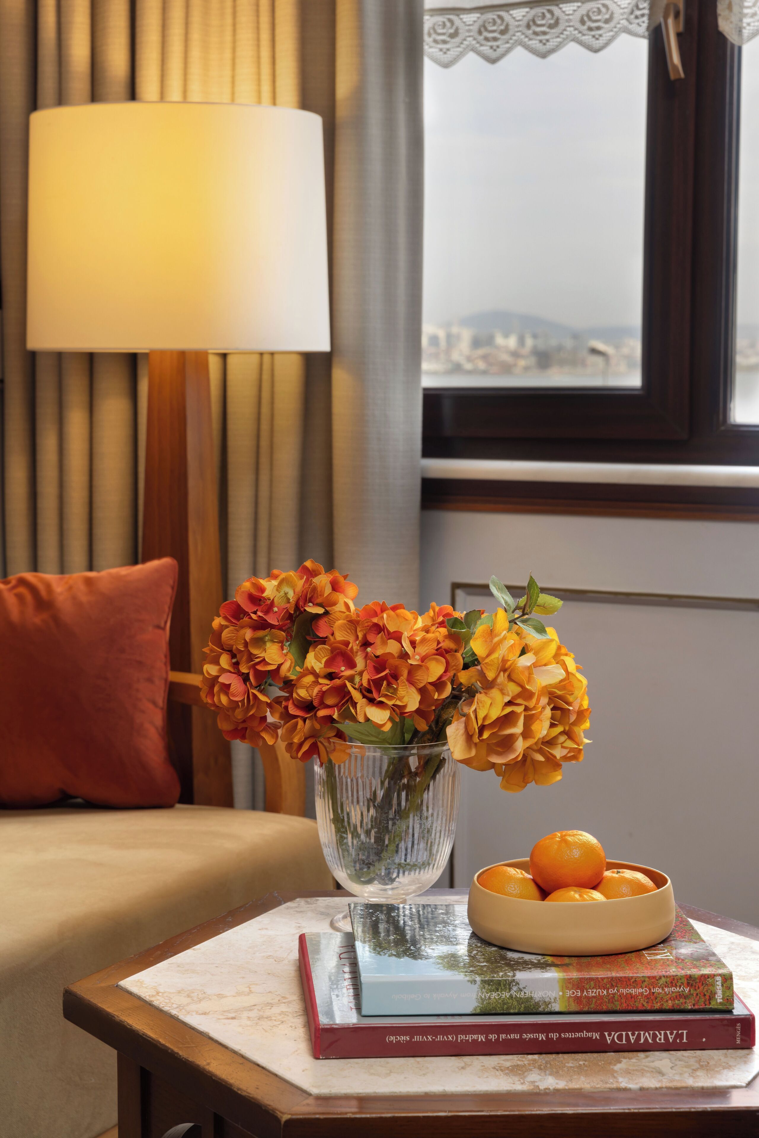 Ein Zimmer mit einem Bett, einem Tisch mit Blumen und einer Schale Orangen, einer Lampe und einem Fenster mit Vorhängen.