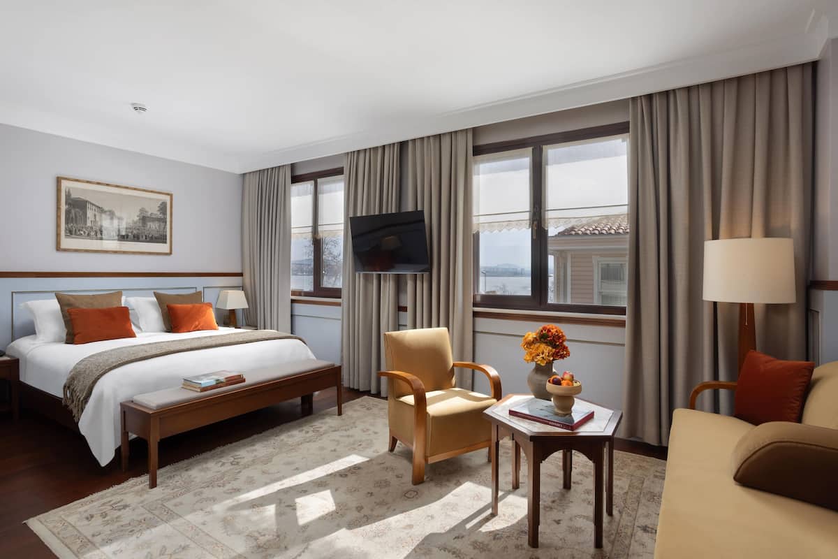 A hotel room with a bed, a chair, a table with a vase, and a TV.