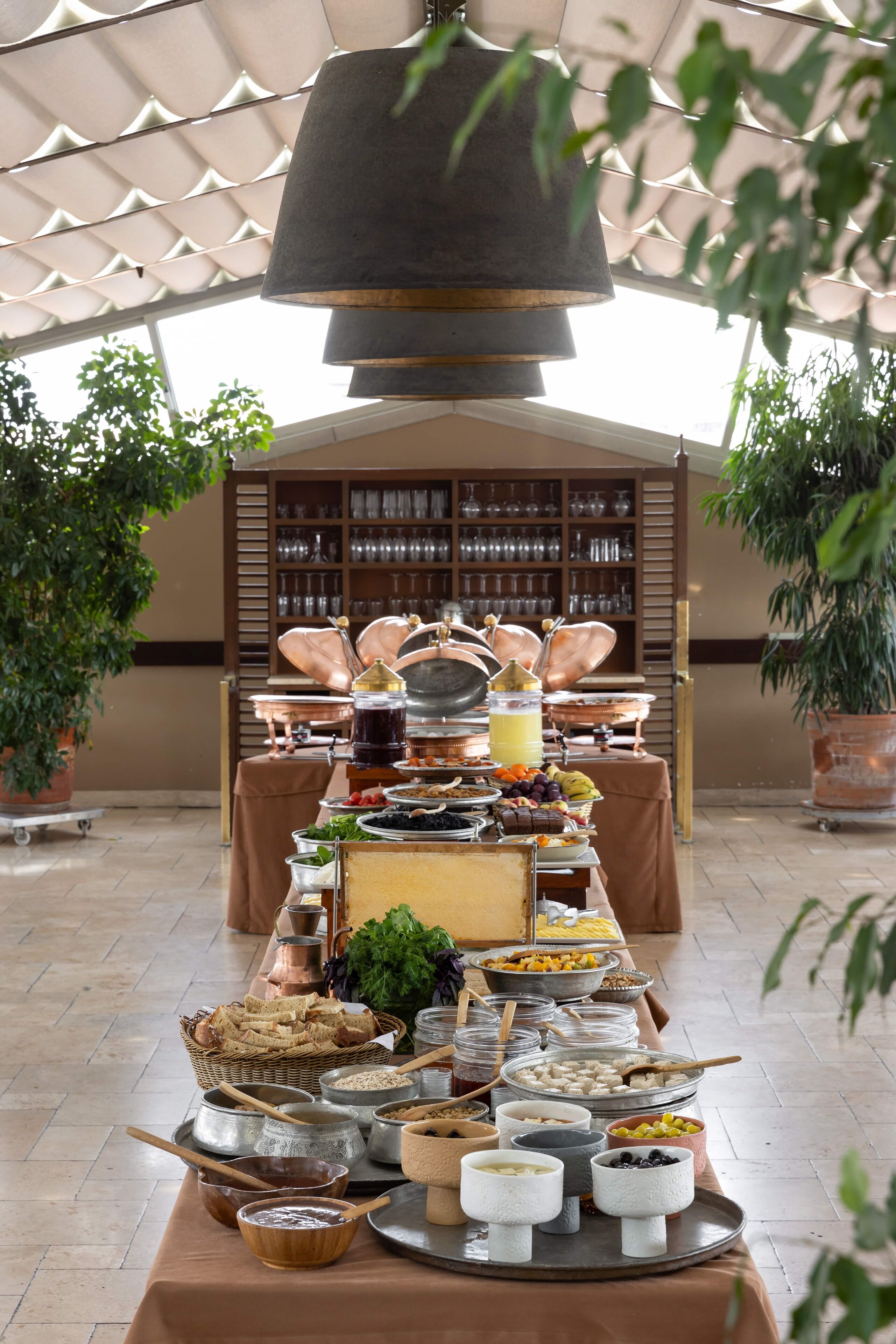 A buffet setup with various food items and utensils on a table, surrounded by potted plants and a backdrop of shelves with glassware.