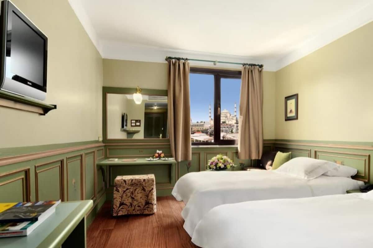A hotel room with two beds, a desk, a television, and a window with a view of a mosque.