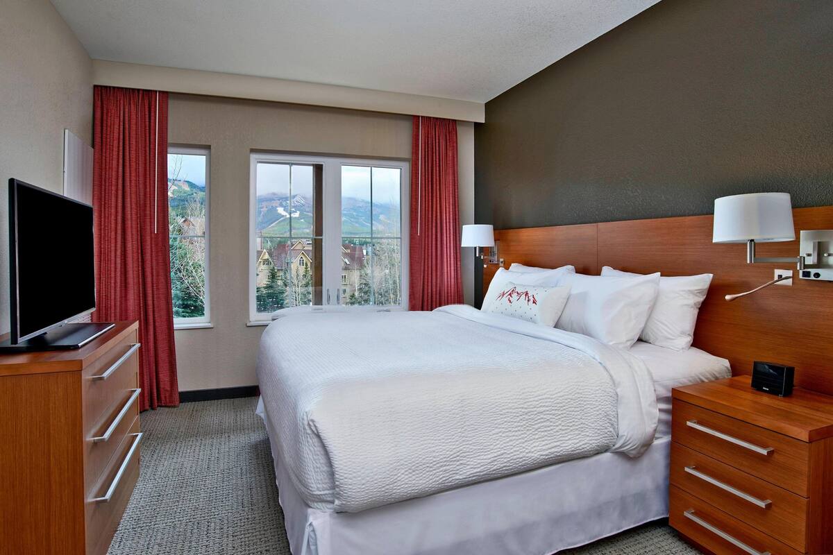 Une chambre d’hôtel avec un grand lit, un téléviseur posé sur une commode en bois et une vue sur les montagnes par les fenêtres.