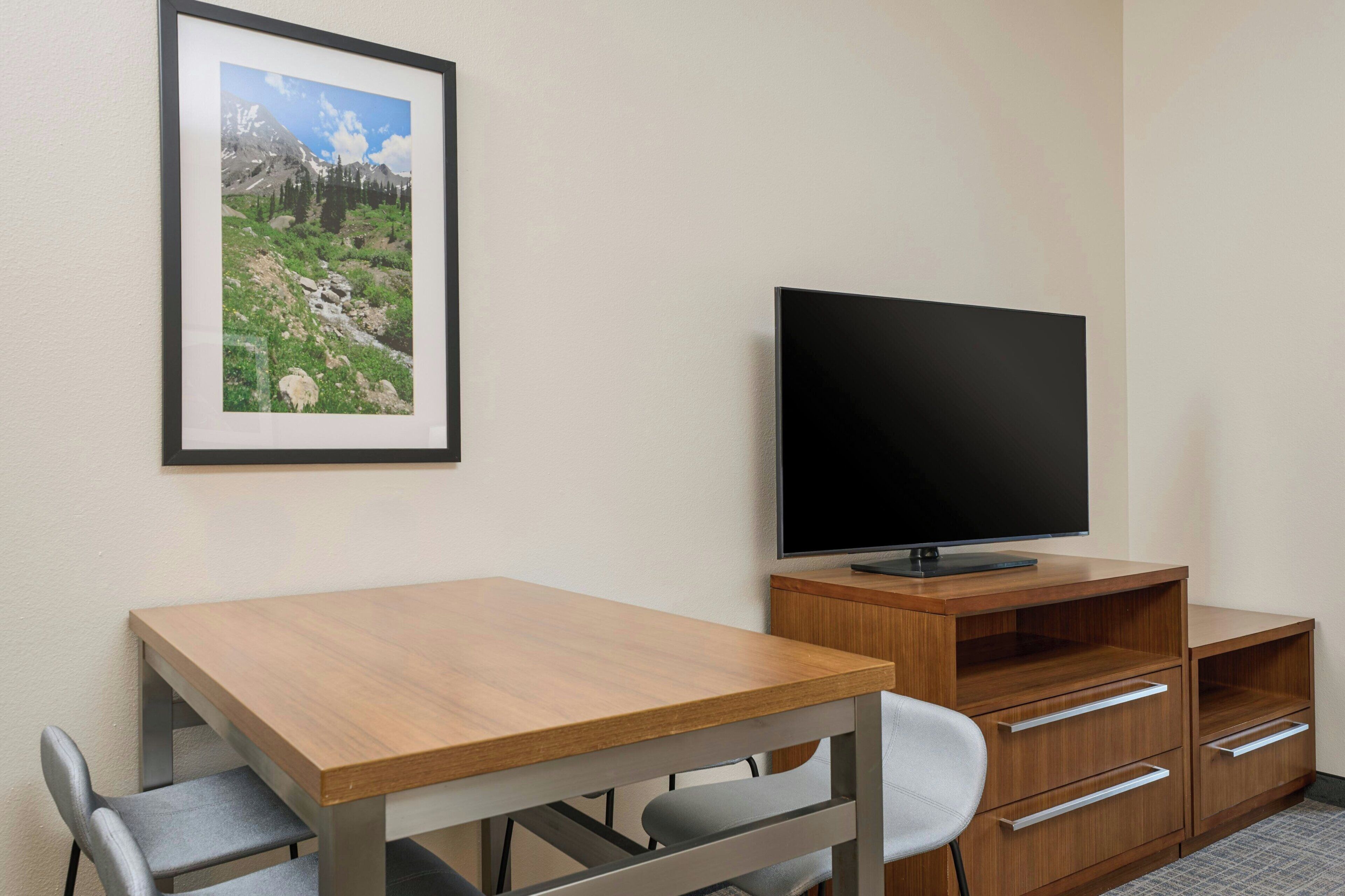 Une chambre d’hôtel avec un bureau en bois, un téléviseur posé sur un meuble en bois, un tableau représentant un paysage de montagne, et une chaise.