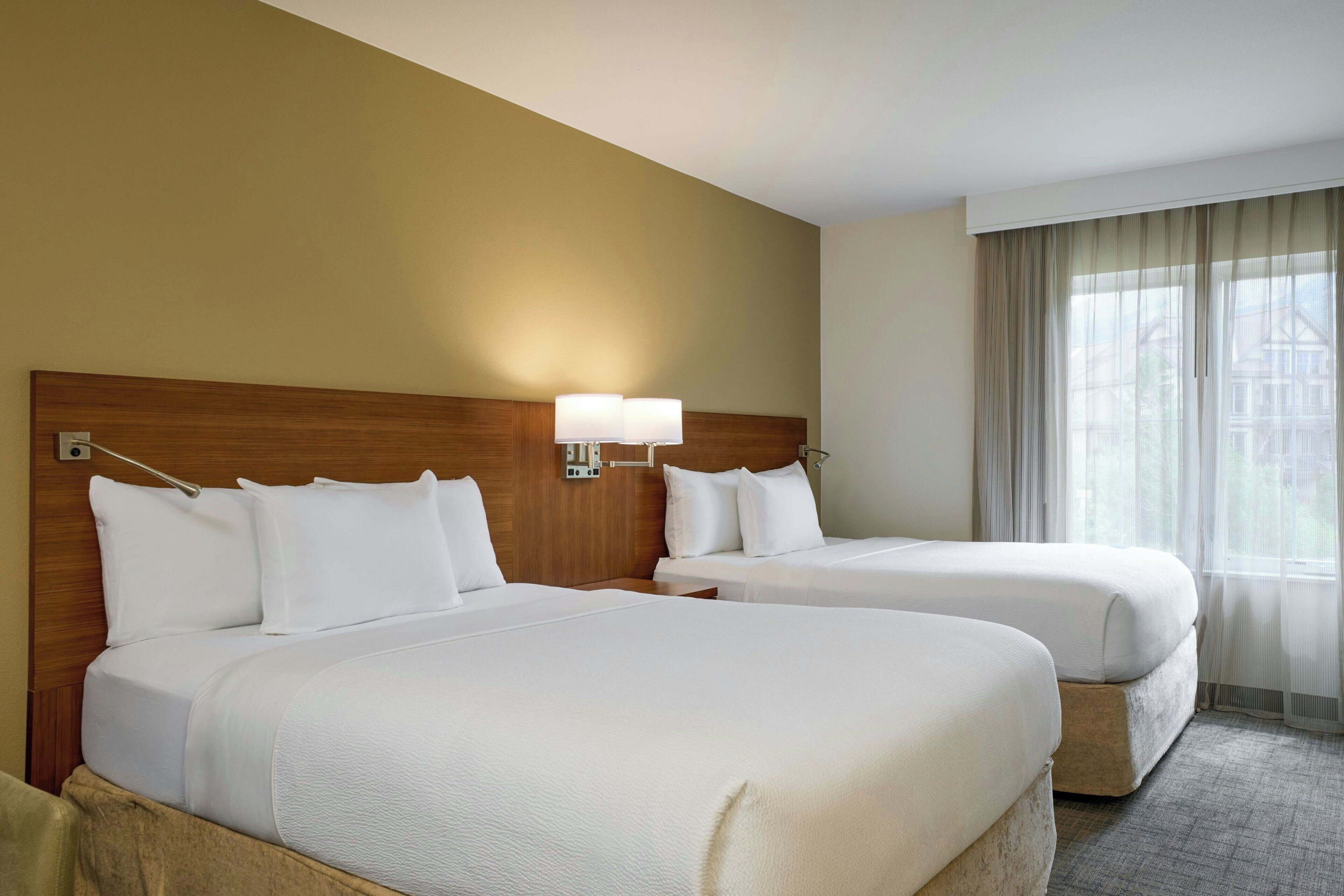 Une chambre d’hôtel avec deux lits, une tête de lit en bois, une table de chevet, une lampe et une fenêtre avec des rideaux.