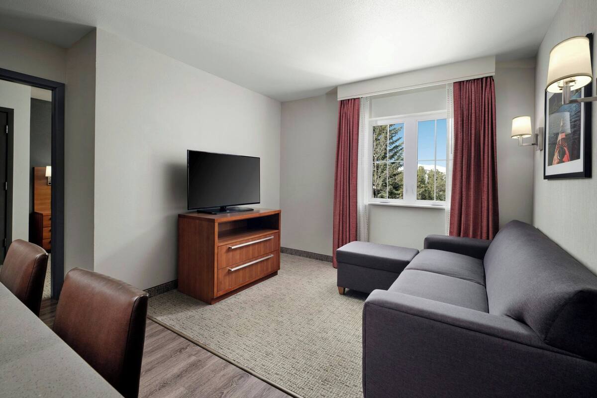 Une chambre d’hôtel moderne dotée d’un téléviseur à écran plat, d’un meuble de rangement en bois, d’un canapé gris et d’une grande fenêtre ornée de rideaux rouges.