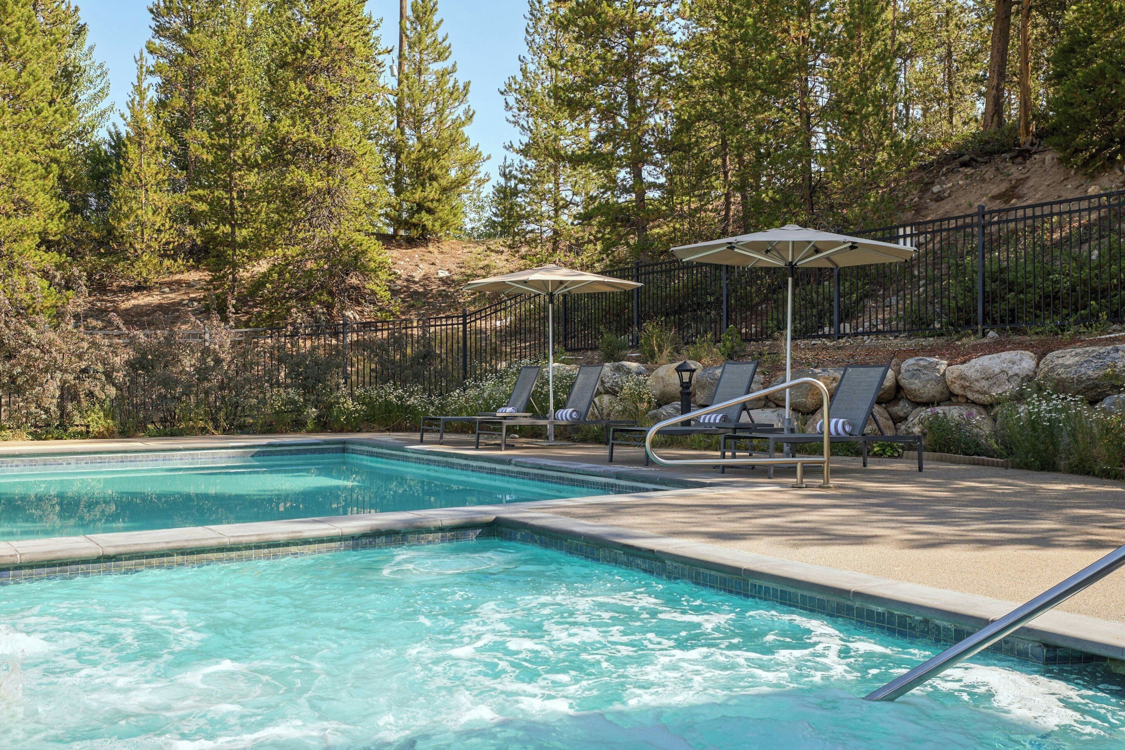 A swimming pool with lounge chairs and umbrellas, surrounded by a fence and trees.