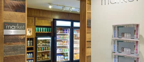 A section of a hotel with wooden paneling, a refrigerated display case with beverages, and a wall with 'The Market' sign and newspapers.