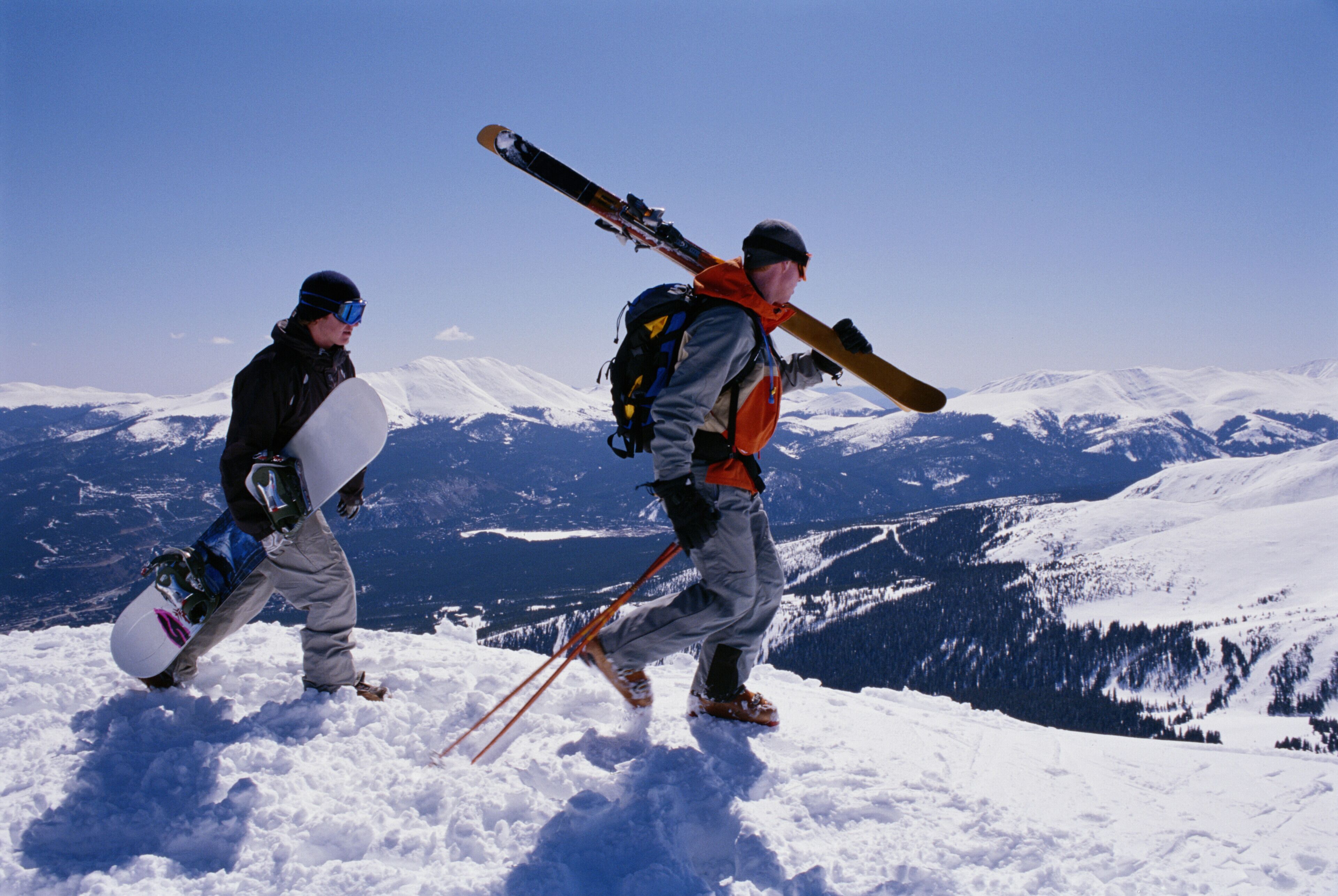 Dois esquiadores caminhando em uma montanha nevada, com pranchas de snowboard e esquis.