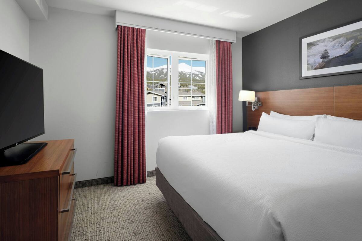 Une chambre d’hôtel avec un grand lit, un téléviseur à écran plat posé sur une commode en bois, une fenêtre avec des rideaux rouges et un tableau encadré accroché au mur.