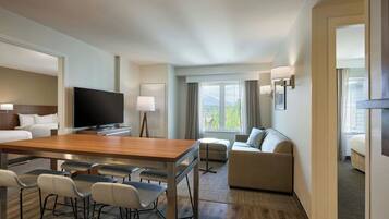 A hotel room with a wooden dining table, bar stools, a sofa, a TV, and a window with curtains.